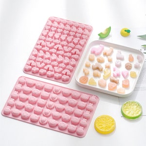 May include: Two pink silicone moulds for making candy or soap. The larger mould has a variety of animal shapes, including cats, dogs, and rainbows. The smaller mould has a variety of animal faces, including pigs, cats, and lions. The moulds are shown with a white plate of colourful candies.
