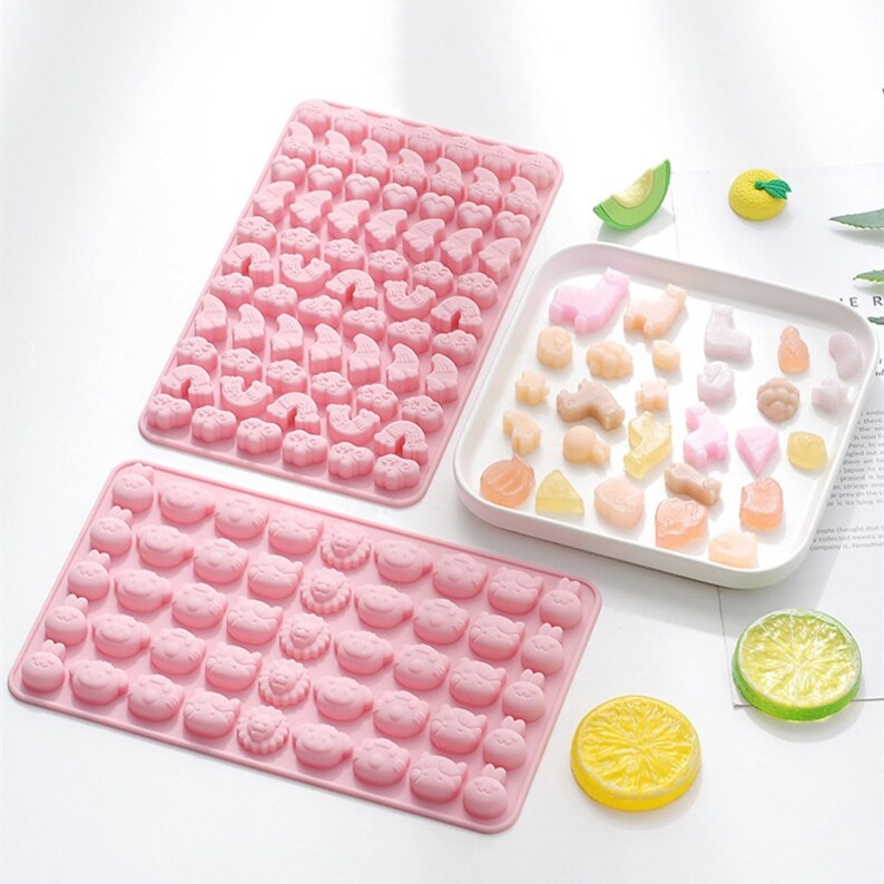 May include: Two pink silicone moulds for making candy or soap. The larger mould has a variety of animal shapes, including cats, dogs, and rainbows. The smaller mould has a variety of animal faces, including pigs, cats, and lions. The moulds are shown with a white plate of colourful candies.