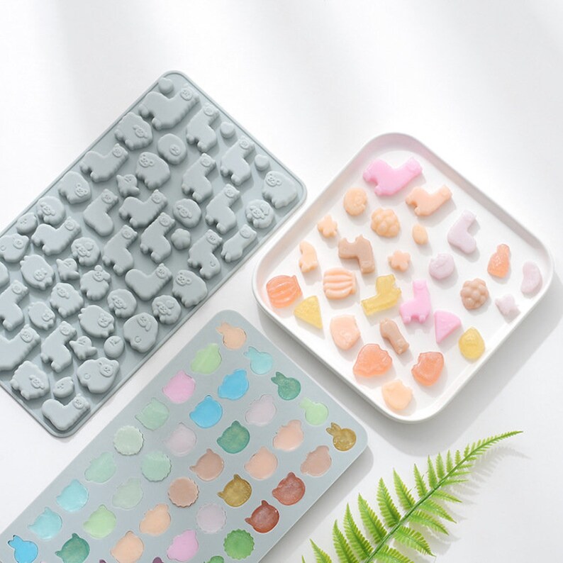May include: Three silicone moulds for making candy or chocolate. The moulds are grey and feature different animal designs. One mould has a repeating pattern of llamas, another has a variety of animals, and the third has a repeating pattern of bunnies. A white plate with a variety of colourful candies in the shapes of the animals is shown.