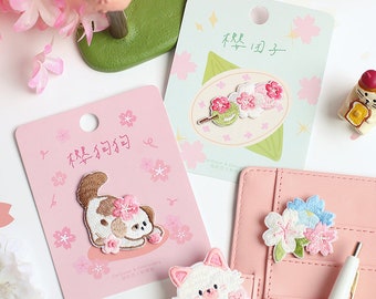 May include: Four embroidered iron-on patches with Japanese text and floral designs. The patches feature a cat, a dog, a flower arrangement, and a skewer of mochi with cherry blossoms. The patches are on pink and white backgrounds.