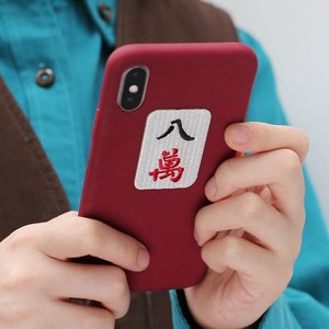 May include: A red phone case with a white square sticker featuring Chinese characters in black and red. The characters are "人" and "場".
