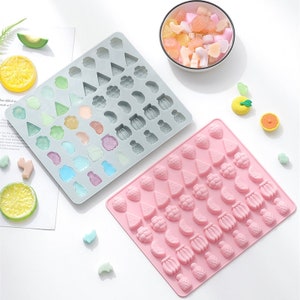 May include: Two silicone moulds for making fruit-shaped sweets. The moulds are grey and pink and feature a variety of fruit shapes, including strawberries, pineapples, bananas, lemons, and watermelons.