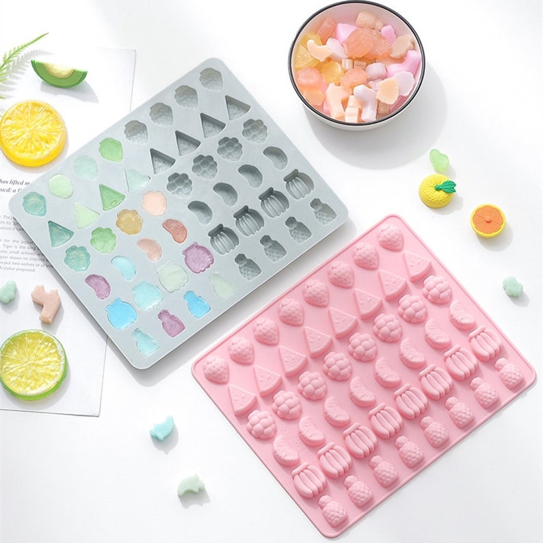 May include: Two silicone moulds for making fruit-shaped sweets. The moulds are grey and pink and feature a variety of fruit shapes, including strawberries, pineapples, bananas, lemons, and watermelons.