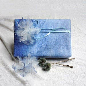 May include: A blue gift box wrapped in blue paper with a white and blue flower on top. The box is tied with a light blue ribbon. There are two small dried flowers on the white surface below the box.