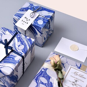 Puede incluir: Tres cajas de regalo envueltas en papel marmoleado azul y blanco con diferentes estilos de cinta y etiqueta. Las etiquetas tienen el texto "Buena suerte" y "Mi amor" con un mensaje sobre bromas pr&aacute;cticas.