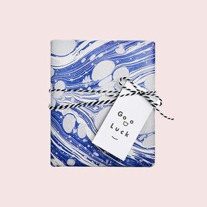 Puede incluir: Papel de regalo marmoleado azul y blanco con una etiqueta de regalo blanca que dice "Good Luck" con una carita sonriente.