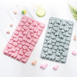 May include: Two silicone moulds for making candy or chocolate. One mould is pink and the other is grey. Both moulds feature a repeating pattern of small llama and pig shapes.