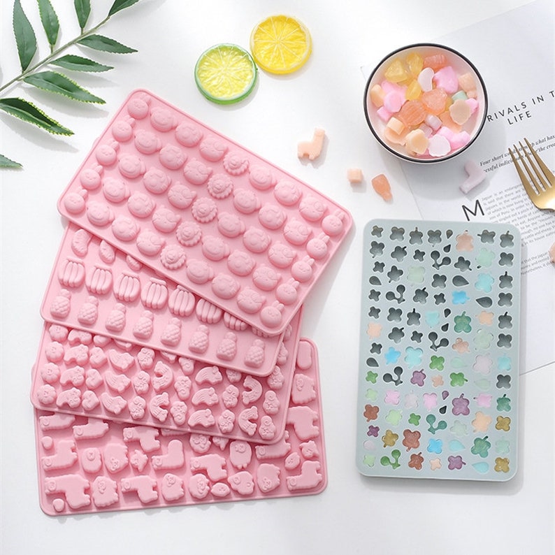 May include: Five pink silicone moulds for making candy or chocolate. The moulds feature different designs, including animals, fruits, and flowers. A grey silicone mould with a variety of small flower and leaf designs is also shown.