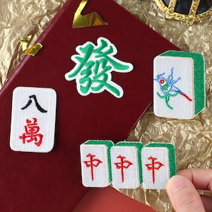 May include: Five embroidered iron-on patches featuring Chinese mahjong tiles. The tiles are white with green borders and red Chinese characters. One tile features a bird with blue and red accents.
