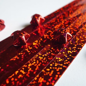 May include: Red iridescent paper strips with red origami stars scattered on a white surface.