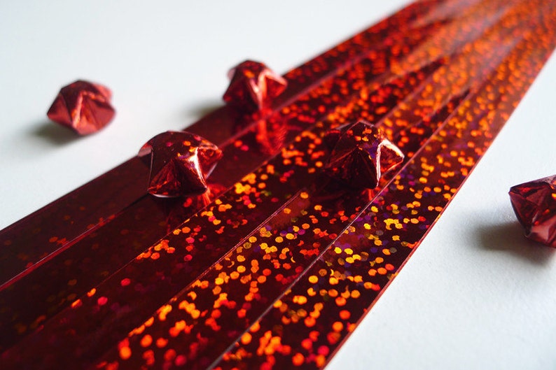 May include: Red iridescent paper strips with red origami stars scattered on a white surface.