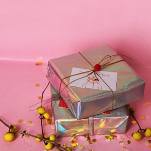 May include: Two iridescent gift boxes wrapped in silver paper with a brown ribbon and a small red bow. The top box has a handwritten card that says "You are the best". The boxes are on a pink surface with gold confetti.