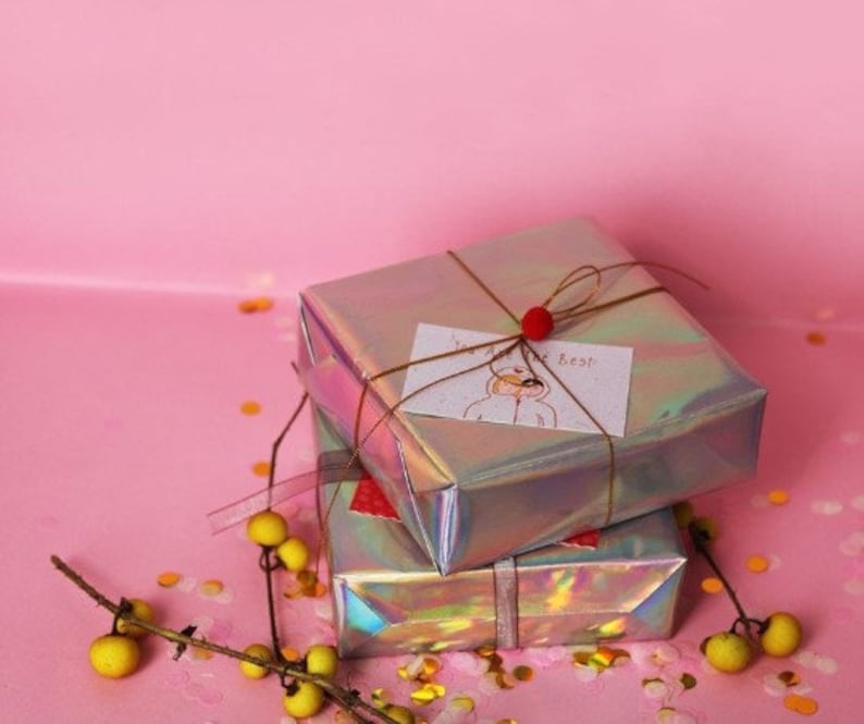 May include: Two iridescent gift boxes wrapped in silver paper with a brown ribbon and a small red bow. The top box has a handwritten card that says "You are the best". The boxes are on a pink surface with gold confetti.
