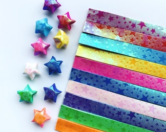 May include: A set of ten colorful origami paper strips with a star pattern. The strips are arranged in a rainbow pattern, with the red strip at the top and the orange strip at the bottom. There are also several origami stars made from the paper, scattered around the strips.