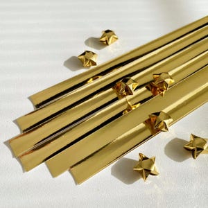 May include: A collection of gold-coloured star-shaped embellishments and strips on a white background. The stars are three-dimensional and folded. The strips are long and rectangular with a reflective surface. These items are likely for crafting or decorative use.