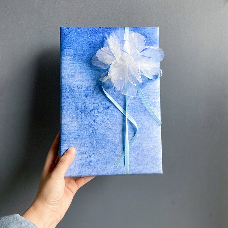 May include: A blue gift box with a white paper flower and blue ribbon. The box has a watercolour-like design.