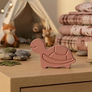 Pode incluir: Um brinquedo em forma de tartaruga rosa claro está sobre uma superfície de madeira. O brinquedo tem um design de casco segmentado. No fundo, há bichos de pelúcia, uma pequena tenda e uma pilha de cobertores. Uma corda de luzes está pendurada.