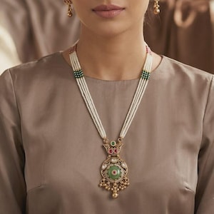 May include: A woman wearing a gold-toned necklace and matching earrings. The necklace features multiple strands of white beads, green and red accents, and a large pendant with a green stone. The earrings have a floral design with red and green accents.