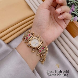 Puede incluir: Un reloj de latón en tono dorado con una correa texturizada y ornamentada, con pequeñas cuentas colgantes y piedras rosas incrustadas. La esfera es redonda con fondo blanco y marcadores simples. El texto Brass High gold Watch No.131.5 es visible.