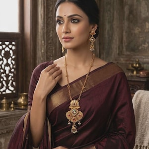 May include: A woman in a maroon sari with gold borders, adorned with a long gold necklace and matching earrings. The necklace features a detailed pendant with colourful gemstones. The image includes the text "A Heritage Story".