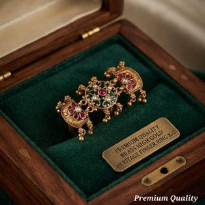 Peut inclure: Une bague en laiton doré ornée d'un motif circulaire central avec émail vert, petites perles blanches et pierres rouges, flanquée de deux panneaux décoratifs avec pierres rouges et perles pendantes. Texte : PREMIUM QUALITY BRASS HIGH GOLD HERITAGE FINGER RING X-2.