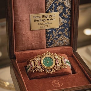 May include: A brass high gold heritage watch with a green face, displayed in a wooden box with a red velvet interior. The watch has a gold band with red and green accents. The box also contains a pink fabric and a blue patterned fabric.
