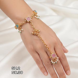 May include: A gold-toned hand harness featuring a chain bracelet connected to a ring. The design includes square pink stones, a blue triangular stone, and small dangling white pearls with green accents. The text SH.2 NO.30 is visible on the side.