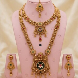 May include: A gold-coloured heritage jewellery set, comprising a necklace, earrings, and a headpiece, displayed on a pink stand. The set features intricate designs with red, green, and white accents. The text "High Gold Heritage Combo Set F-19" is visible.