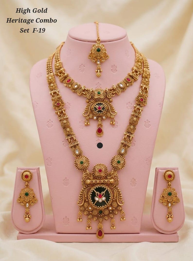 May include: A gold-coloured heritage jewellery set, comprising a necklace, earrings, and a headpiece, displayed on a pink stand. The set features intricate designs with red, green, and white accents. The text "High Gold Heritage Combo Set F-19" is visible.