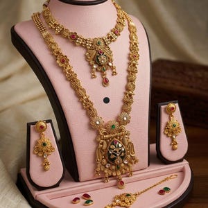 May include: A gold-coloured jewellery set, including a necklace, earrings, and a headpiece, displayed on a pink stand. The necklace and earrings feature floral and ornate designs with red, green, and white accents. The text "High Gold Heritage Combo Set F-18" is visible.