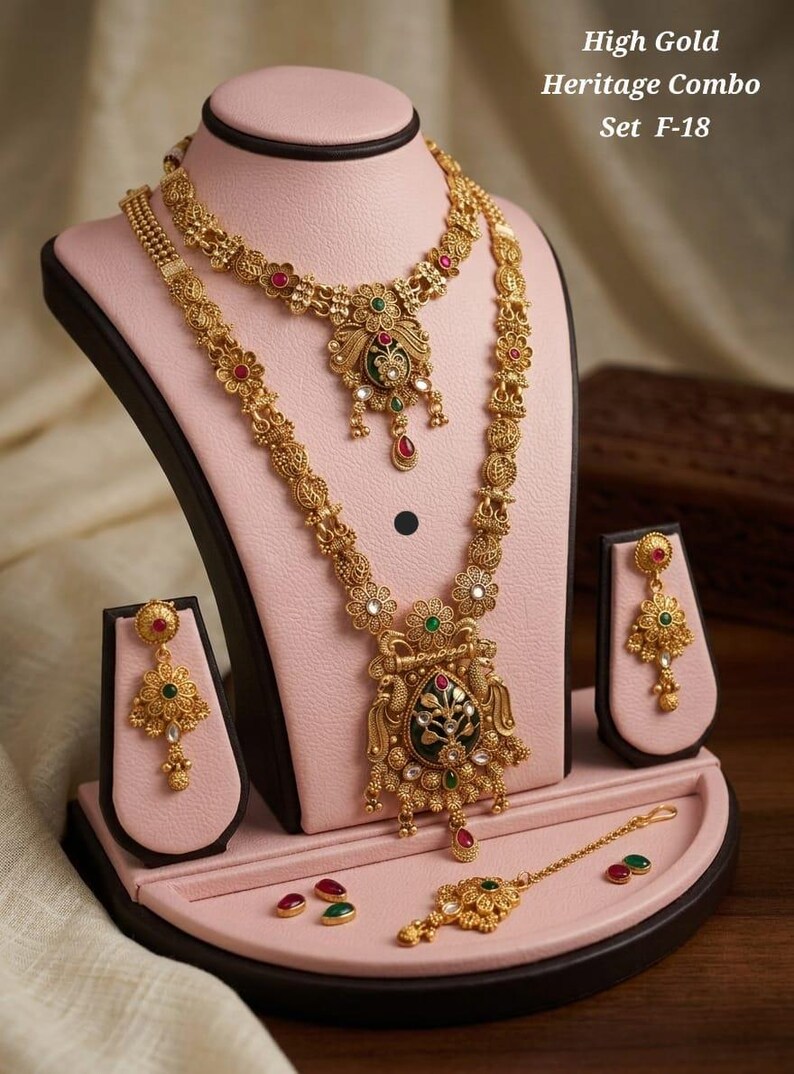 May include: A gold-coloured jewellery set, including a necklace, earrings, and a headpiece, displayed on a pink stand. The necklace and earrings feature floral and ornate designs with red, green, and white accents. The text "High Gold Heritage Combo Set F-18" is visible.