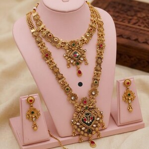 May include: A gold-coloured heritage jewellery set, comprising a necklace, earrings, and a headpiece, presented on a pink stand. The necklace showcases intricate floral and geometric designs with red, green, and white accents. The set is labelled "High Gold Heritage Combo Set F-17."