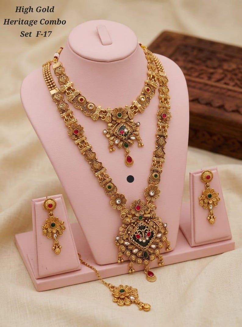 May include: A gold-coloured heritage jewellery set, comprising a necklace, earrings, and a headpiece, presented on a pink stand. The necklace showcases intricate floral and geometric designs with red, green, and white accents. The set is labelled "High Gold Heritage Combo Set F-17."