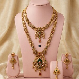 May include: A gold-coloured heritage jewellery set, comprising two necklaces, earrings, and a headpiece, presented on pink stands. The necklaces showcase intricate floral and geometric designs with red, green, and gold accents. The text "High Gold Heritage Combo Set F-16" is visible.