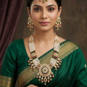 May include: An ornate gold-toned necklace, earrings, and headpiece set. The necklace features multiple strands of white beads, interspersed with green and red accents, and a large pendant with a central green stone. The jewelry is set against a green sari.