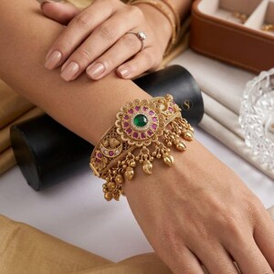 May include: A gold-toned cuff bracelet featuring a central floral motif with a green stone, surrounded by small pink stones and clear crystals. The band has intricate metalwork, crescent moon accents, and dangling gold charms along the bottom edge.