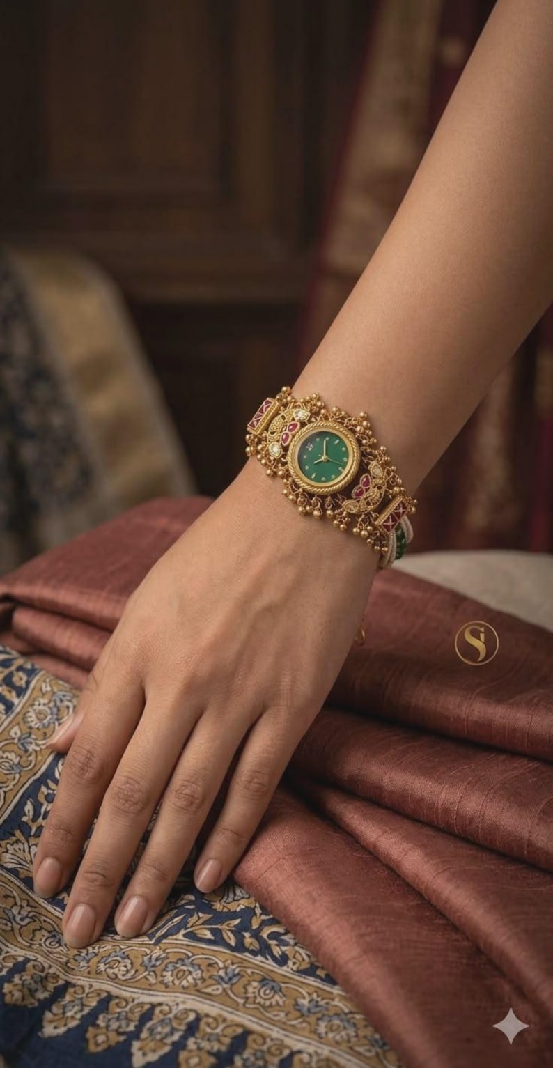 May include: Gold-toned wristwatch with a green face and ornate detailing. The watch features red and white accents and is worn on a wrist. The background includes a patterned fabric.
