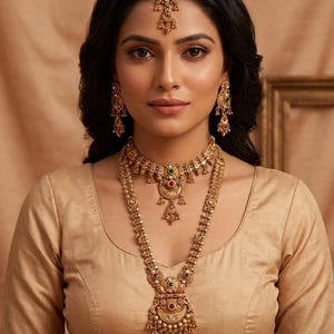 May include: A gold-toned jewelry set featuring a long necklace with a large ornate pendant, a matching choker necklace, dangling earrings, and a maang tikka headpiece. Each piece is detailed with small beads and embedded red and green stones.