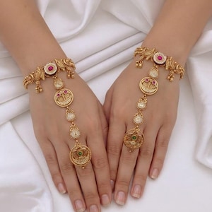 May include: A pair of gold-toned hand harnesses featuring intricate metalwork, circular medallions, and small dangling charms. The jewellery includes embedded pink and green stones, connecting a wrist bracelet to a ring with a central chain. Text: SH.Tas.Q.10 Barsh.kopr NO.80