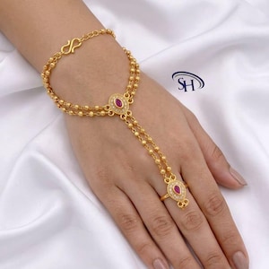 May include: A gold-tone hand harness featuring a double-strand beaded chain connecting a wrist bracelet to a finger ring. Both the central oval centerpiece and the teardrop-shaped ring component are set with a small red stone and clear crystals.