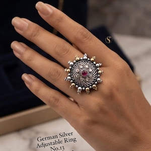May include: An adjustable German silver ring featuring a circular, sunburst-inspired design with oxidized silver swirls, small gold and silver beads along the outer edge, and a central round red stone. Text reads: German Silver Adjustable Ring No.13 D.No.00055.