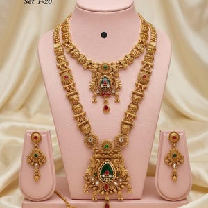 May include: A gold-coloured heritage jewellery set, comprising a necklace, earrings, and a headpiece, presented on a pink stand. The set showcases detailed craftsmanship with red, green, and white embellishments. The text "High Gold Heritage Combo Set F-20" is visible.