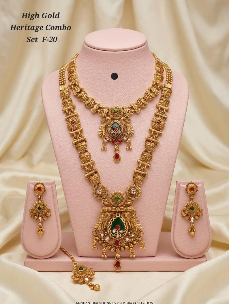 May include: A gold-coloured heritage jewellery set, comprising a necklace, earrings, and a headpiece, presented on a pink stand. The set showcases detailed craftsmanship with red, green, and white embellishments. The text "High Gold Heritage Combo Set F-20" is visible.