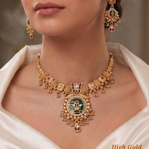 Peut inclure: Un ensemble collier et boucles d'oreilles doré avec un pendentif circulaire central orné d'un émail vert, de motifs floraux et de petites pierres rouges. Le bijou comporte des perles dorées et blanches, avec le texte High Gold Heritage Set D-81.