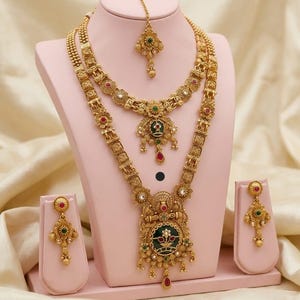 May include: A gold-coloured heritage jewellery set, including a necklace, earrings, and a headpiece, displayed on a pink stand. The set features intricate floral and geometric designs with red, green, and white accents. The text "High Gold Heritage Combo Set F-21" is visible.