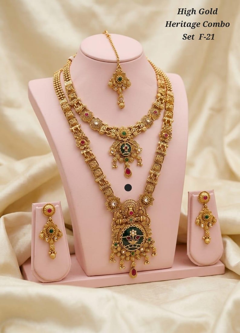 May include: A gold-coloured heritage jewellery set, including a necklace, earrings, and a headpiece, displayed on a pink stand. The set features intricate floral and geometric designs with red, green, and white accents. The text "High Gold Heritage Combo Set F-21" is visible.