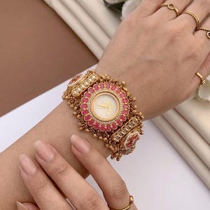 May include: Ornate gold-coloured watch with a circular face and a floral pattern. The watch has a pink and white bezel, and is decorated with small, round details. The watch is worn on a wrist, with a neutral-toned background.