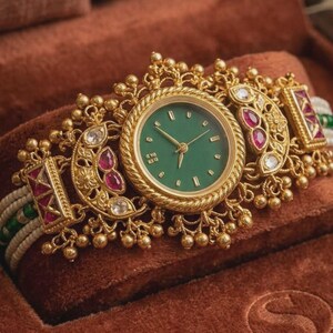 May include: Ornate gold-toned wristwatch with a green watch face. The watch features intricate detailing, including red and white gemstones, and is set on a white and green beaded band. The watch is displayed in a brown velvet box.
