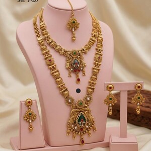 May include: A gold-coloured heritage jewellery set, including a necklace, earrings, and headpiece, displayed on a pink mannequin. The necklace features intricate detailing with red, green, and clear stone accents. The text "High Gold Heritage Combo Set F-20" is visible.