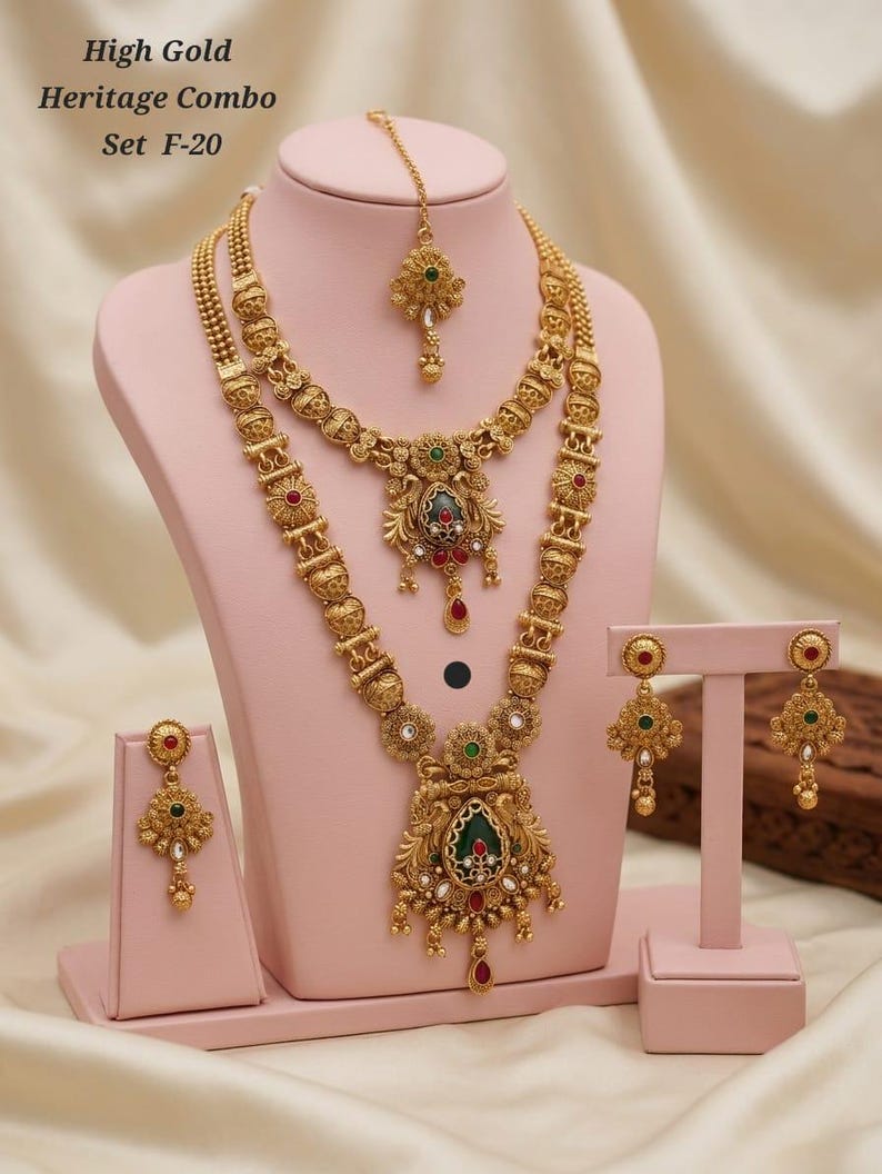 May include: A gold-coloured heritage jewellery set, including a necklace, earrings, and headpiece, displayed on a pink mannequin. The necklace features intricate detailing with red, green, and clear stone accents. The text "High Gold Heritage Combo Set F-20" is visible.